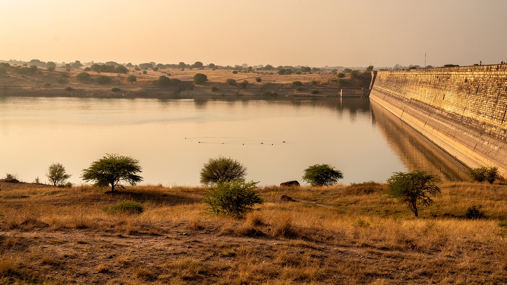 Calm water of Dholidhaja Dam with a stone wall and flat grassland at golden hour